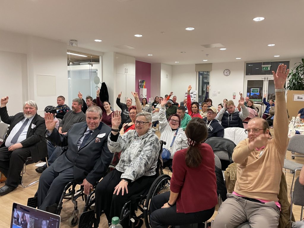 Image of members at the 2024 AGM sitting down, grinning and raising their arms in celebration